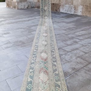 Peut inclure: Un tapis long et étroit avec un fond crème et des motifs complexes en vert, rose et rouge. Le tapis est plié en deux et posé sur un sol en carrelage gris.