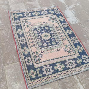May include: A rectangular rug with a central pink field and a dark blue border. The border features geometric patterns and floral motifs in green and white. The rug has a red fringe along the edges, adding a decorative touch.