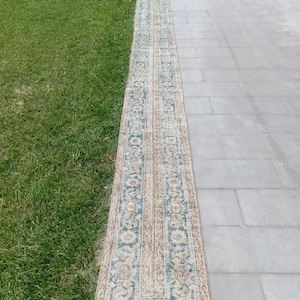 Könnte beinhalten: Ein langer, schmaler Läufer mit einem sich wiederholenden Blumenmuster in Blau- und Beigetönen. Der Teppich hat ein Mittelteil mit komplexen botanischen Motiven und eine dekorative Bordüre, die eine abgenutzte Textur aufweist.