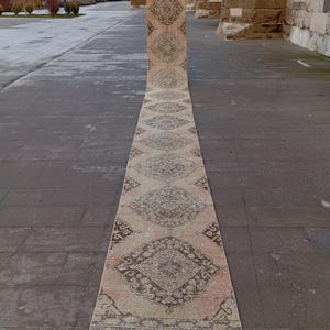 May include: A long, narrow, vintage runner rug with a repeating geometric pattern in shades of beige, brown, and faded rose. The rug is displayed outdoors, showcasing its length and intricate design. The rug's edges are slightly frayed, adding to its antique charm.