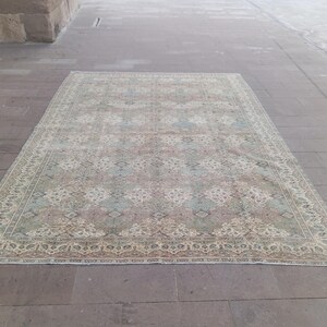 May include: Rectangular area rug with a faded, vintage appearance. The rug features a central pattern in muted shades of pink, blue, and beige, framed by a detailed border. The rug is placed on a stone surface.