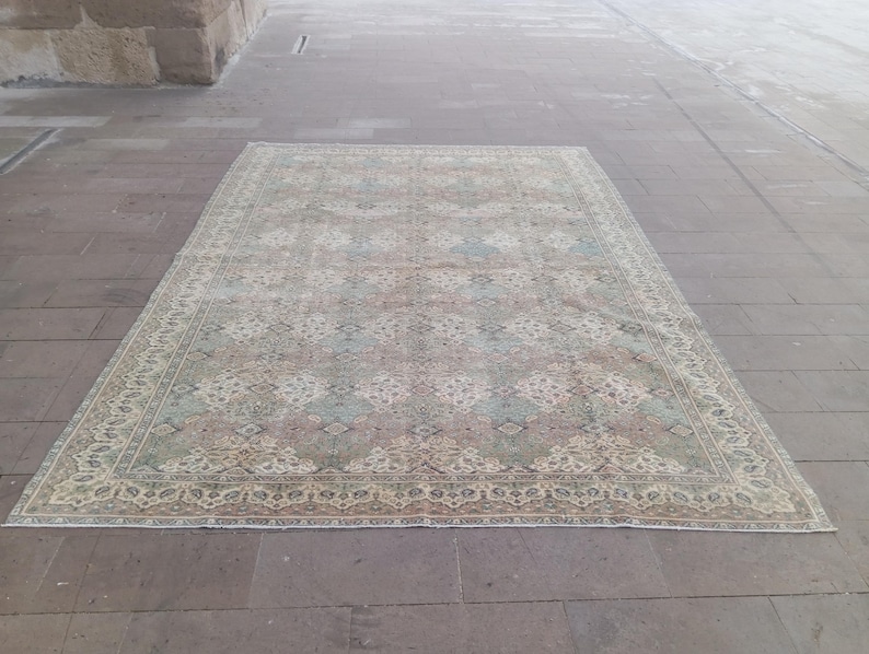 May include: Rectangular area rug with a faded, vintage appearance. The rug features a central pattern in muted shades of pink, blue, and beige, framed by a detailed border. The rug is placed on a stone surface.