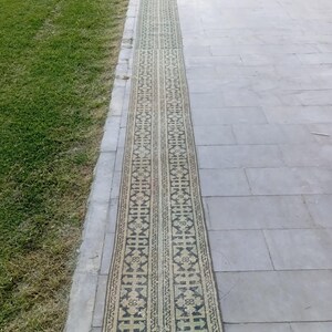 May include: A long, narrow rug with a repeating geometric pattern in shades of beige, green, and black. The rug is rectangular and appears to be made of woven material. It is placed on a grey stone surface next to a patch of green grass.