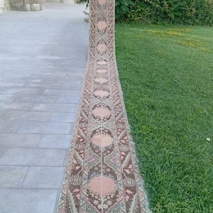 May include: A long, narrow runner rug featuring a repeating geometric pattern in shades of dusty pink, brown, and muted green. The rug displays a series of interconnected hexagonal medallions along its length, finished with a short fringe at the end.