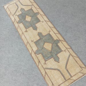 May include: A rectangular beige rug with a geometric design. The rug features a central pattern of two symmetrical teal-coloured shapes with diamond-shaped centres. Brown lines outline the teal shapes and border the rug.