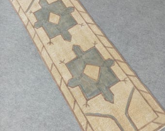May include: A rectangular beige rug with a geometric design. The rug features a central pattern of two symmetrical teal-colored shapes with diamond-shaped centers. Brown lines outline the teal shapes and border the rug.