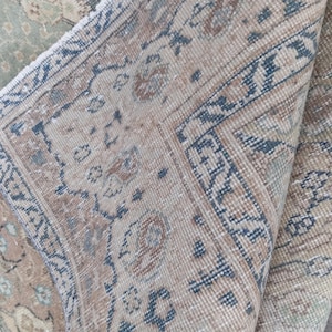May include: A section of a vintage rug with a folded edge, showcasing intricate patterns in beige, blue, and muted rose tones. The rug's design features floral and geometric motifs, creating a classic, aged aesthetic. The rug's texture appears soft and worn.