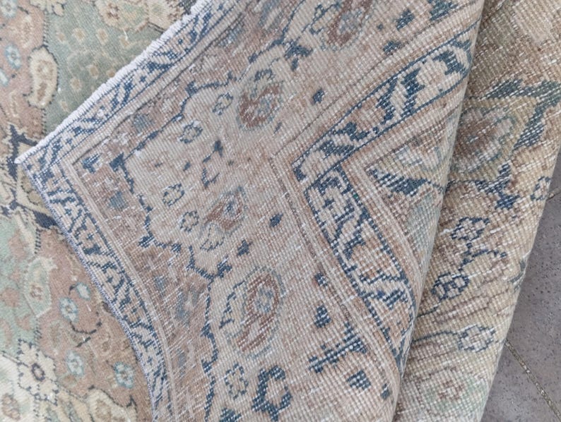 May include: A section of a vintage rug with a folded edge, showcasing intricate patterns in beige, blue, and muted rose tones. The rug's design features floral and geometric motifs, creating a classic, aged aesthetic. The rug's texture appears soft and worn.