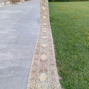 May include: A long, narrow runner rug featuring a faded, intricate pattern in shades of beige, brown, muted red, and soft blue. The rug displays a repeating geometric and floral motif throughout its length, with a worn, vintage-inspired texture.