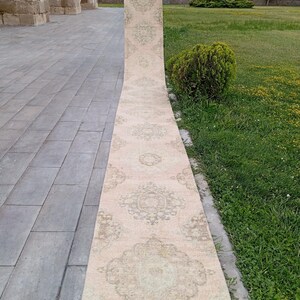 May include: A long, narrow runner rug featuring a pale beige base color with repeating intricate floral and medallion patterns in muted sage green and soft brown tones. The rug displays a symmetrical design throughout its entire length.