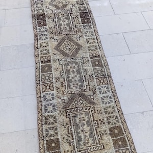 Op de afbeelding: Een lang, rechthoekig tapijt met een beige achtergrond en bruine geometrische patronen. Het tapijt heeft een franje rand.