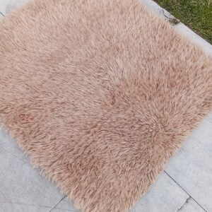 Peut inclure: Un tapis carré et moelleux de couleur marron clair. Le tapis est composé de longues fibres douces, créant un aspect texturé. Le tapis est posé sur une surface carrelée, avec un bout d'herbe verte visible en arrière-plan.