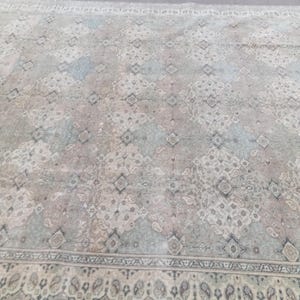 May include: A large, rectangular rug with a faded, vintage appearance. The rug features a repeating geometric pattern in shades of gray, blue, and pink. A detailed border with a paisley design frames the central field. The rug appears to be made of a soft material.