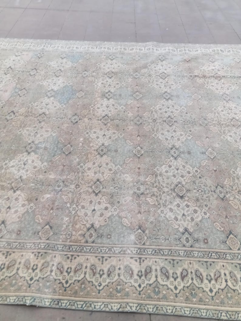 May include: A large, rectangular rug with a faded, vintage appearance. The rug features a repeating geometric pattern in shades of gray, blue, and pink. A detailed border with a paisley design frames the central field. The rug appears to be made of a soft material.
