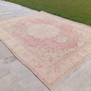 May include: A large rectangular area rug featuring a faded pink and cream colour palette with an intricate floral medallion pattern in the centre and a detailed border design. The rug has a low pile texture and a symmetrical, ornate woven motif.