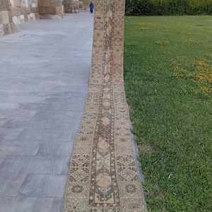 May include: A long, narrow runner rug featuring a faded, intricate geometric pattern in shades of beige, brown, and muted green. The rug has a central medallion design flanked by decorative borders, with a short, light-colored fringe at the end.