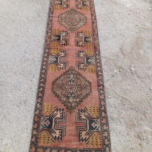 May include: A long, rectangular runner rug featuring a faded salmon-pink background with intricate geometric patterns in shades of brown, mustard yellow, and dark blue. The design includes central diamond motifs and a dark, patterned border.