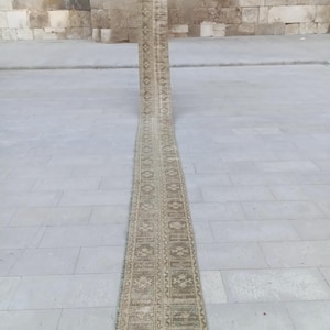 May include: A long, narrow rug with a repeating geometric pattern in beige, brown, and green. The rug is laid out on a light grey stone surface, with a textured stone wall in the background. The rug appears to be vintage or antique, with a faded appearance.