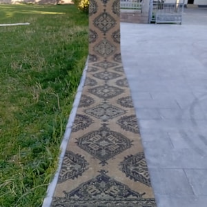 May include: A long, narrow rug with an intricate geometric pattern in shades of beige and dark green. The rug is displayed outdoors, partially rolled out on a concrete surface, showcasing its length and design. The rug's design features repeating diamond and floral motifs.