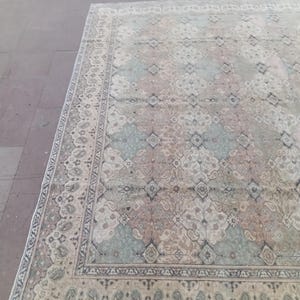 May include: A large, rectangular rug with an intricate, faded floral pattern. The rug features a cream-colored background with accents of blue, pink, and brown. The border has a detailed design in darker shades. The rug is placed on a stone surface.