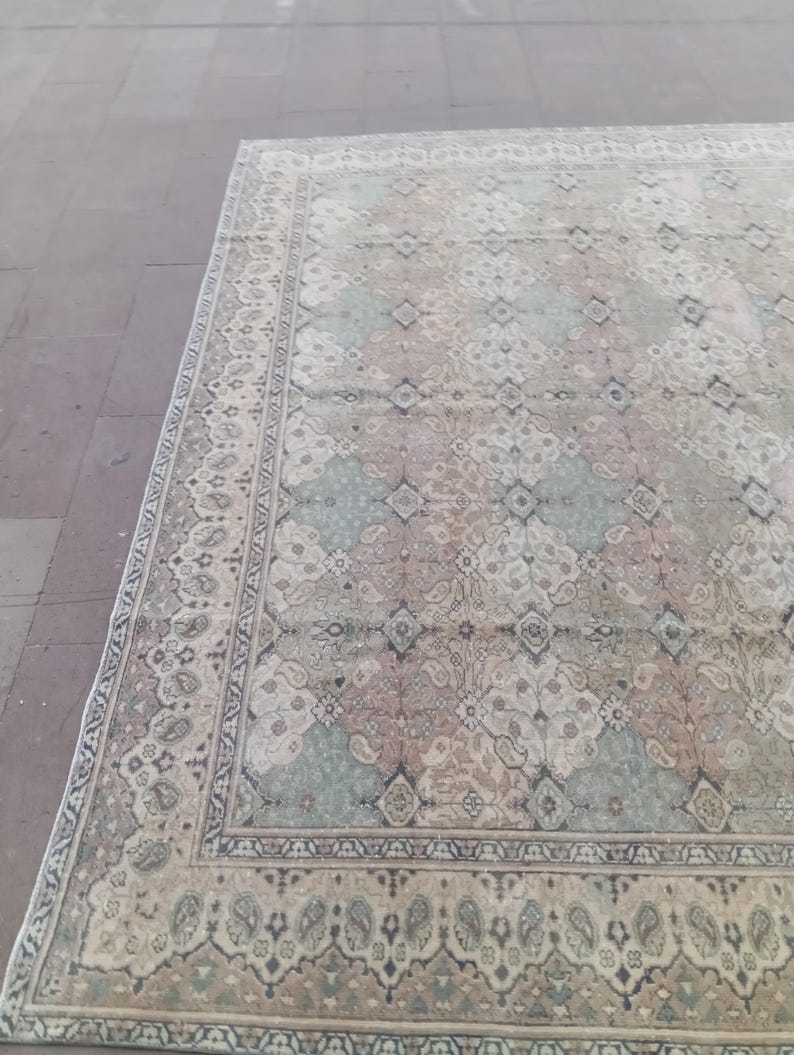 May include: A large, rectangular rug with an intricate, faded floral pattern. The rug features a cream-colored background with accents of blue, pink, and brown. The border has a detailed design in darker shades. The rug is placed on a stone surface.