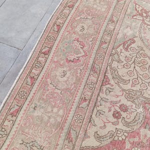May include: A rectangular area rug featuring a detailed woven pattern in shades of muted pink, beige, and soft sage green. The design includes a wide decorative border with repeating geometric and floral motifs surrounding a central field.