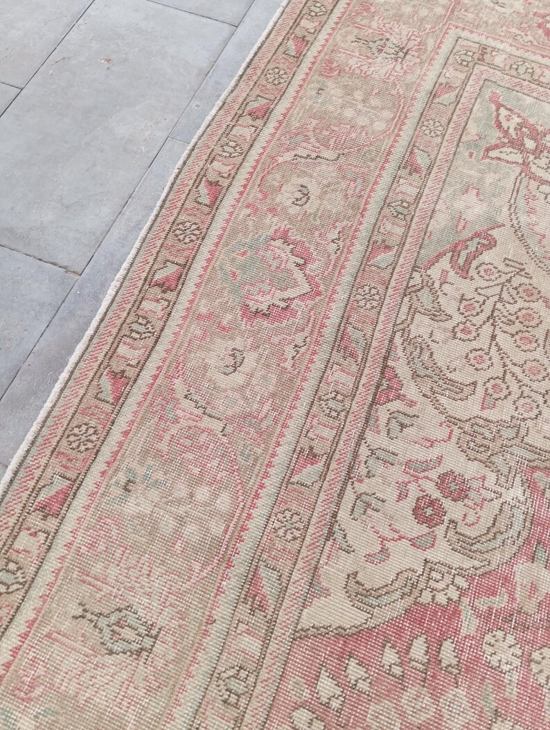 May include: A rectangular area rug featuring a detailed woven pattern in shades of muted pink, beige, and soft sage green. The design includes a wide decorative border with repeating geometric and floral motifs surrounding a central field.