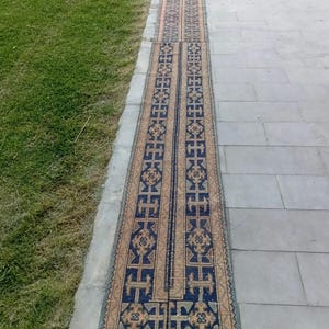 May include: A long, narrow rug with a repeating geometric pattern in navy blue, tan, and light blue. The rug is placed on a gray stone surface next to a patch of green grass.