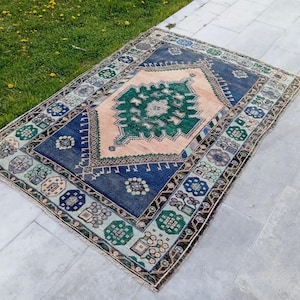 May include: Rectangular rug with a central peach-colored field and a detailed green and blue geometric design. The rug has a dark blue border with floral and geometric patterns in green, blue, and beige. The rug is placed on a gray stone surface.