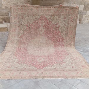 May include: A large rectangular area rug featuring a faded pink and beige colour palette with an intricate central medallion and detailed floral border patterns. The rug displays a distressed, worn texture throughout its woven surface design.