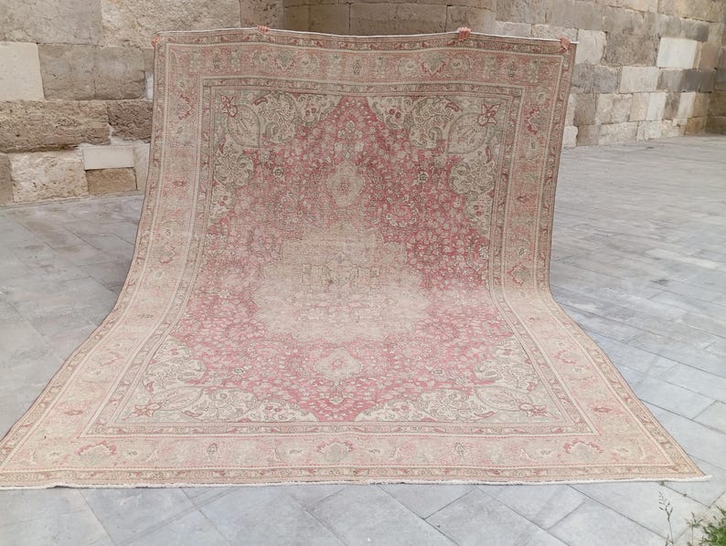 May include: A large rectangular area rug featuring a faded pink and beige colour palette with an intricate central medallion and detailed floral border patterns. The rug displays a distressed, worn texture throughout its woven surface design.