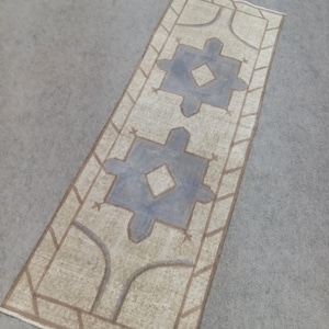 Puede incluir: Alfombra rectangular beige con diseño geométrico. La alfombra presenta dos grandes formas azul claro con centros de diamante, enmarcadas por un borde marrón. La alfombra está colocada sobre una superficie gris.