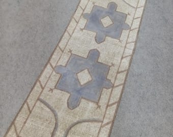May include: Rectangular beige rug with a geometric design. The rug features two large, light blue shapes with diamond centers, framed by a brown border. The rug is placed on a gray surface.