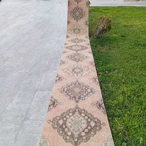 May include: A long, narrow runner rug featuring a faded peach-coloured base with a repeating series of dark brown and muted green geometric medallion patterns. The rug has a flat-woven texture and a fringed edge at the bottom end of the piece.