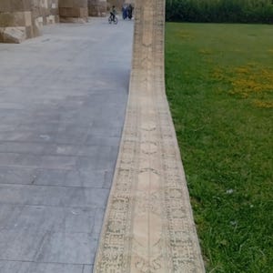 Könnte beinhalten: Ein außergewöhnlich langer und schmaler Läuferteppich in verblassten Beige- und Hellgrüntönen. Der Teppich zeigt ein sich wiederholendes geometrisches und florales Muster über die gesamte Länge sowie ein markantes Bordüren- und Medaillondesign.