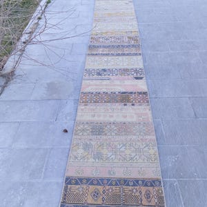 May include: A long, rectangular rug with a patchwork design. The rug features a variety of colors, including pink, blue, yellow, and beige. The rug has a geometric pattern and is likely made of woven fabric.