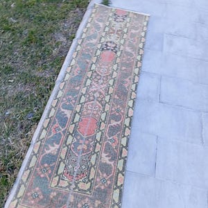 May include: A rectangular, patterned rug with a central design featuring red, pink, and black elements. The rug has a border with green and yellow accents. The rug is placed on a gray stone surface next to a patch of green grass.