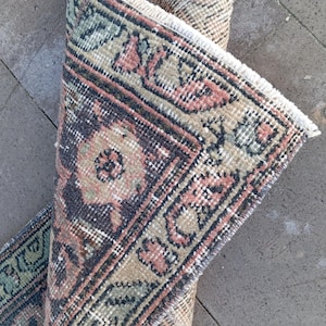 May include: Rolled-up, vintage-style rug with a detailed, geometric pattern. The rug features a color palette of muted tones, including brown, pink, green, and navy blue. The edges are finished with a fringe. This rug is suitable for home decor.