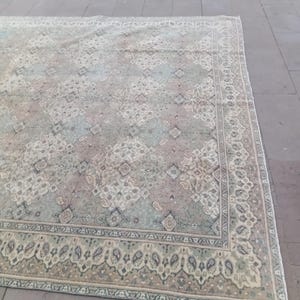 May include: A large, rectangular rug with an intricate, faded floral pattern in muted tones of green, beige, and mauve. The rug features a detailed border with paisley and geometric designs, creating a vintage aesthetic. The rug is placed on a stone surface.