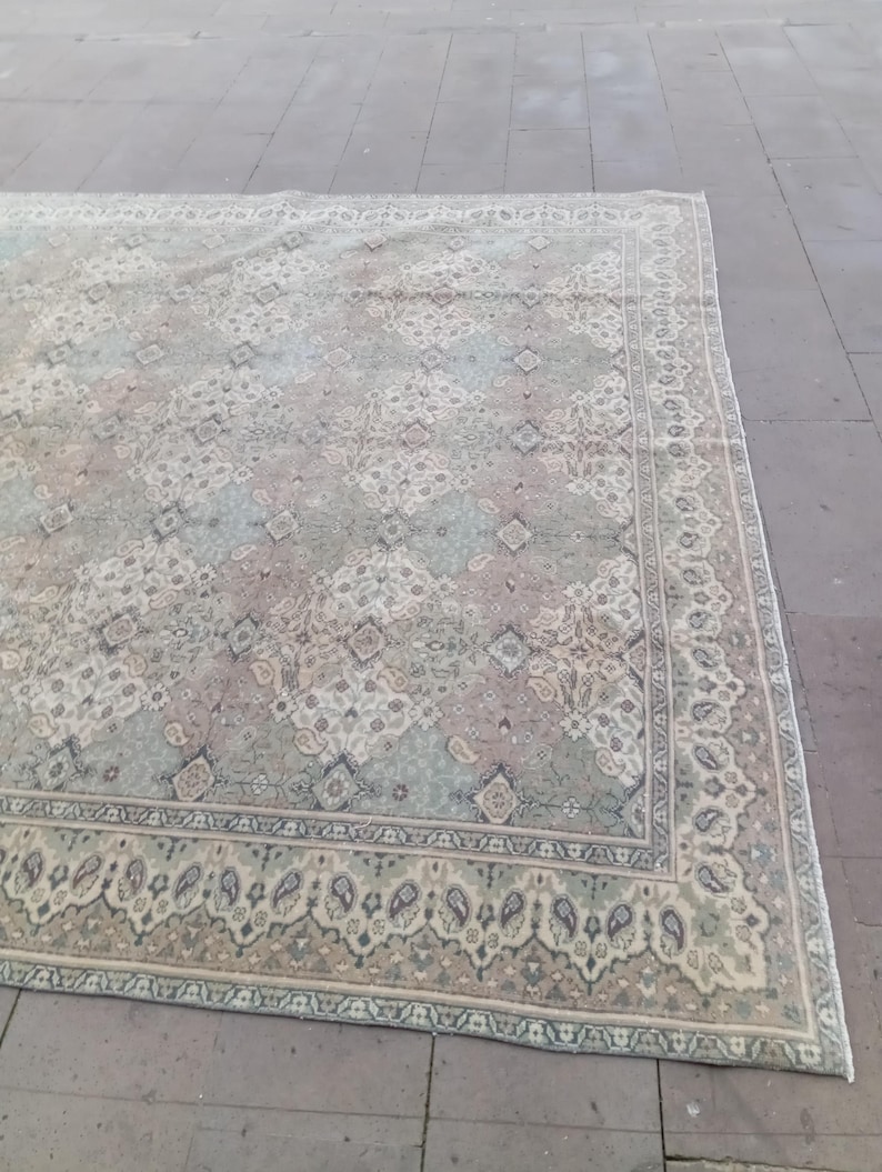 May include: A large, rectangular rug with an intricate, faded floral pattern in muted tones of green, beige, and mauve. The rug features a detailed border with paisley and geometric designs, creating a vintage aesthetic. The rug is placed on a stone surface.