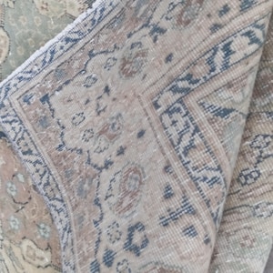May include: Close-up of a section of a vintage rug with a folded corner. The rug features a detailed floral pattern in muted tones of beige, blue, and green. The border has a geometric design in blue and brown.