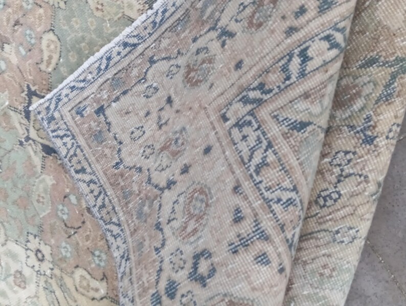 May include: Close-up of a section of a vintage rug with a folded corner. The rug features a detailed floral pattern in muted tones of beige, blue, and green. The border has a geometric design in blue and brown.