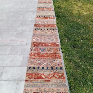 Könnte beinhalten: Ein langer, schmaler Vintage-Teppich mit Patchwork-Design. Der Teppich weist eine Vielzahl von Farben auf, darunter Rot, Orange, Beige und Blau, mit geometrischen und floralen Mustern. Der Teppich wird im Freien auf einer Betonoberfläche präsentiert.
