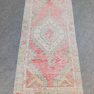 May include: A rectangular rug with a distressed, vintage appearance. The rug features a central diamond pattern in shades of pink and white, framed by a border of light green and beige. Fringes are visible at the ends, adding to its aged aesthetic.