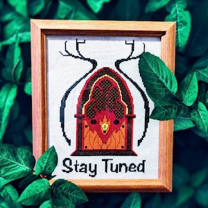 May include: A framed cross-stitch design featuring a vintage radio with antlers and the text "Stay Tuned". The radio is red, orange, and yellow, and the background is white.