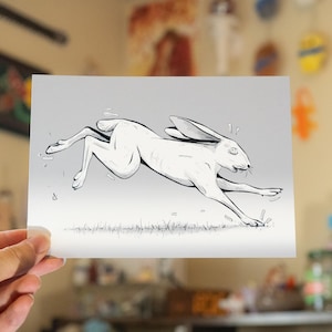 May include: A monochrome illustration of a running hare. The artwork shows the hare mid-jump, with its ears back and legs outstretched. The style is minimalist, using fine lines and shading to create depth and movement. The hare is set against a grey background.