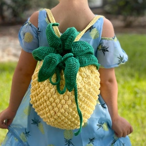 May include: A young child wearing a blue dress with a pineapple pattern is wearing a crocheted pineapple backpack. The backpack is yellow with green leaves.