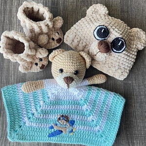 Könnte beinhalten: Eine gehäkelte Babydecke mit einem hellblauen und weißen Streifenmuster. Ein kleiner gehäkelter Teddybär mit braunen Augen und einer braunen Nase sitzt auf der Decke. Die Decke hat ein kleines Flugzeugdesign in der Mitte.