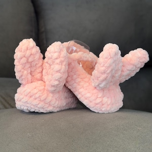 Puede incluir: Pantuflas de conejito rosa con textura peluda. Las pantuflas tienen un pequeño lazo en la parte delantera y están hechas de material suave.