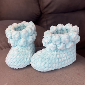 Pode incluir: Um par de botinhas de bebê de tricô azul claro com textura felpuda. As botinhas têm uma bainha dobrada e uma ponta arredondada.